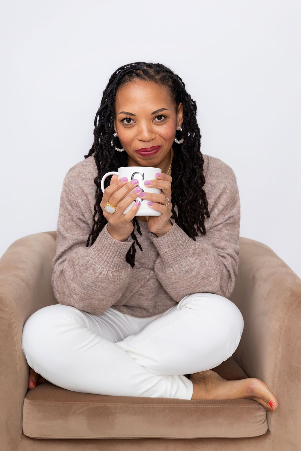 A person in a beige sweater sits cross-legged on a tan armchair while holding a coffee mug.