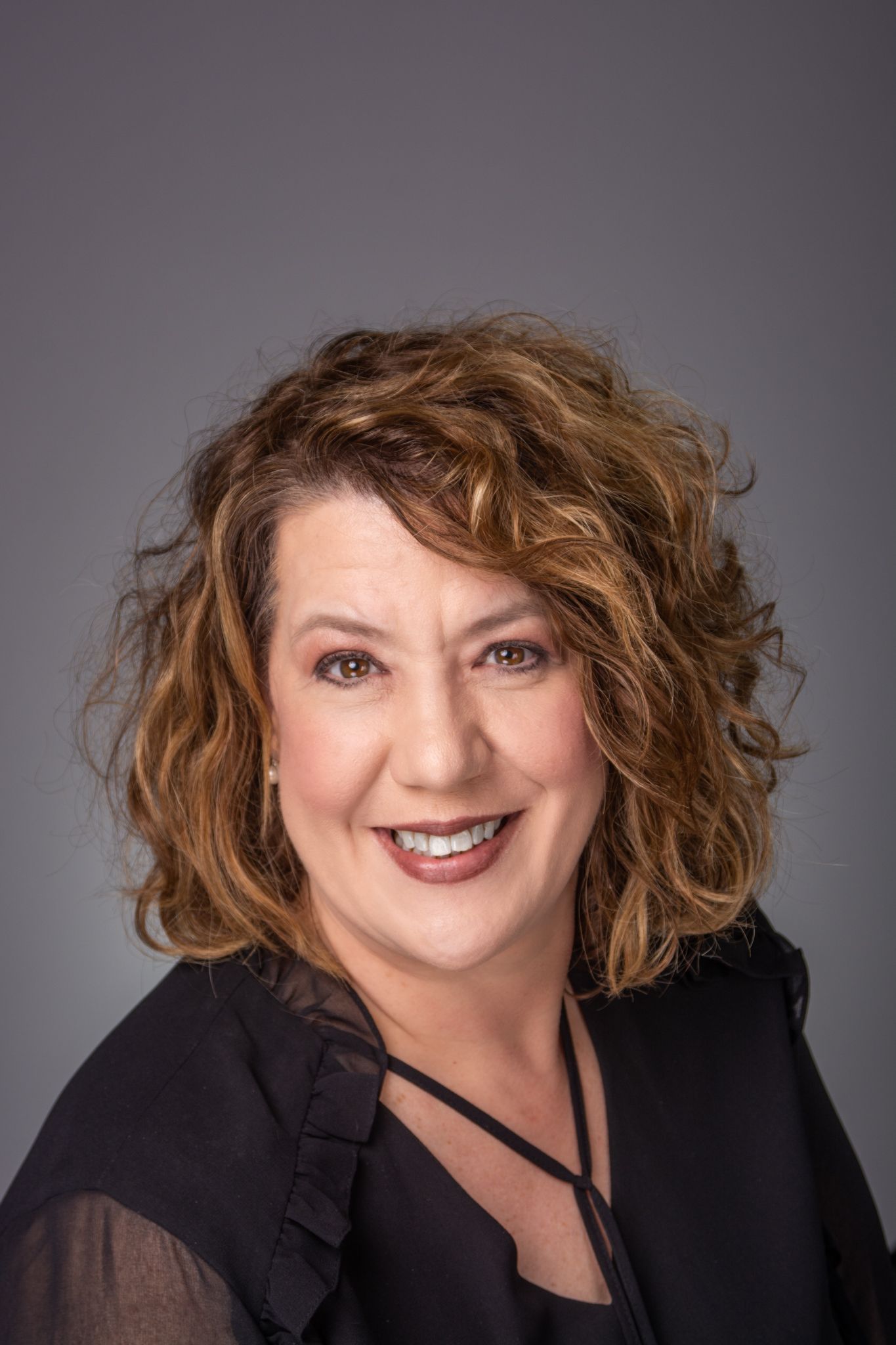 Professional headshot of woman over 50 in a black shirt taken in studio with grey backdrop.