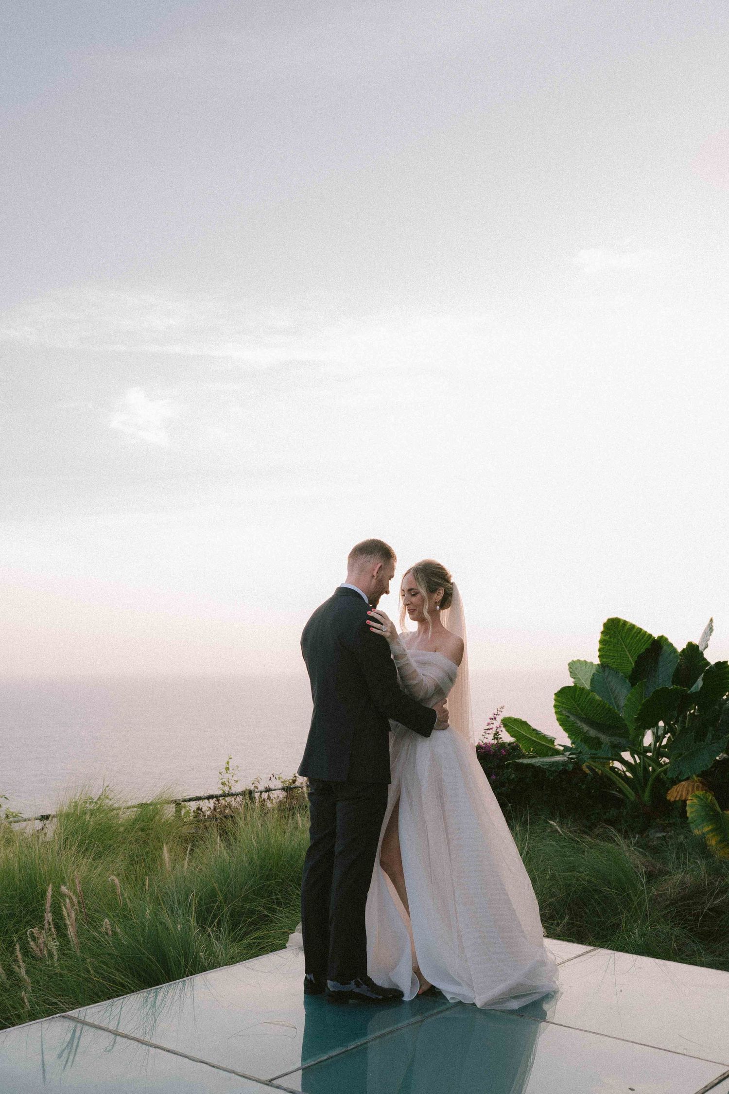 A couple in formal wedding attire shares an intimate moment at sunset on a scenic outdoor terrace with tropical plants.