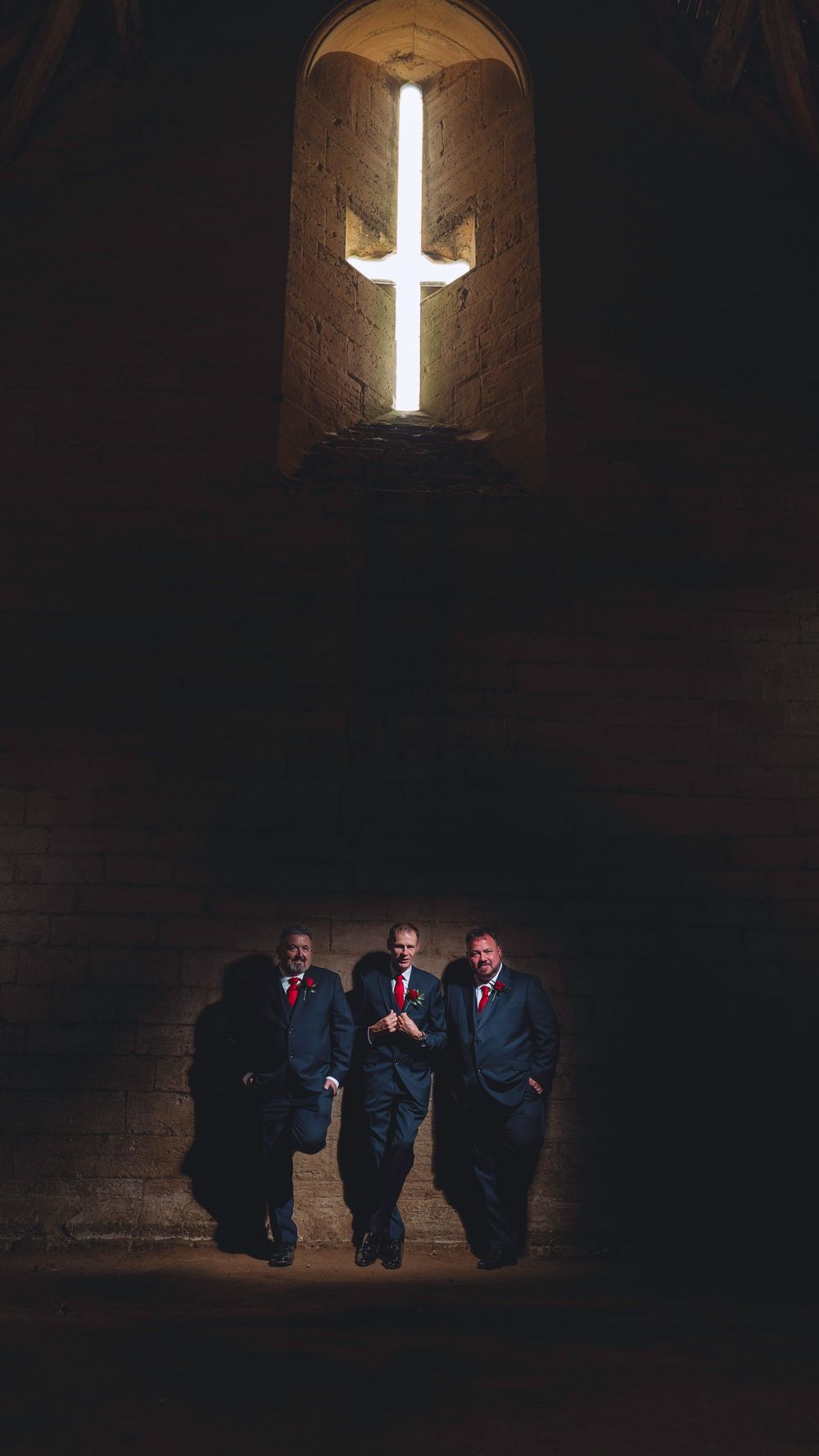 Three figures in dark suits sit beneath a glowing cross in a dimly lit stone church interior.