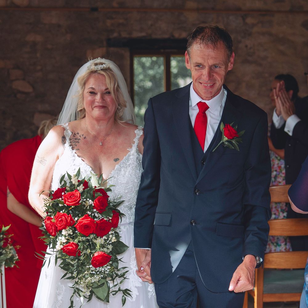 A couple in wedding attire walks down a rustic brick-lined aisle with red roses in the bouquet.