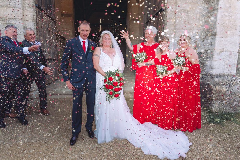 Wedding party exits church while guests shower them with red and white flower petals in celebration.