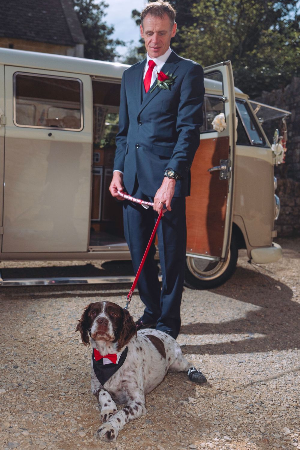 A person in a navy suit stands with a German Shorthaired Pointer dog on a gravel path next to a vintage van.