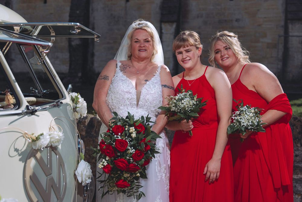 Wedding party wearing red and white dresses pose together holding bouquets at an outdoor celebration.