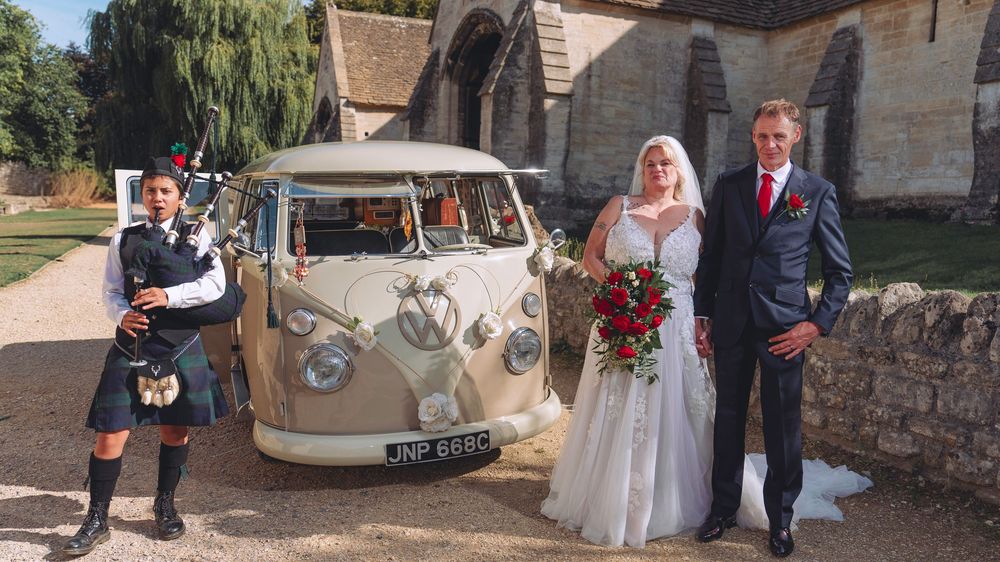Vintage cream-colored classic car decorated with flowers parked outside a rustic stone church on a sunny day.