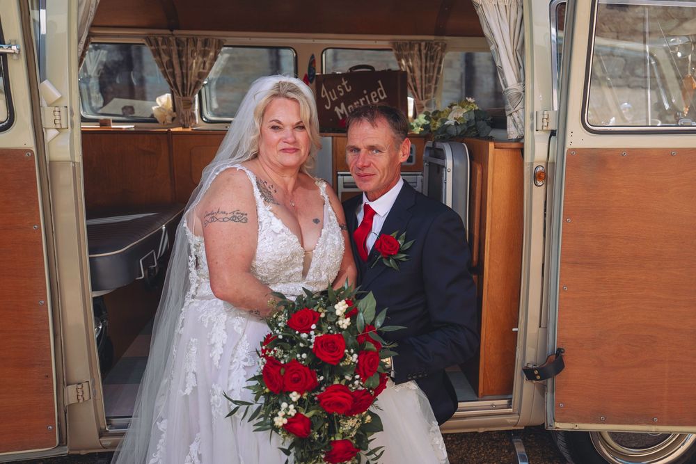 A wedding party poses near vintage orange and cream colored VW camper vans with red rose bouquets.
