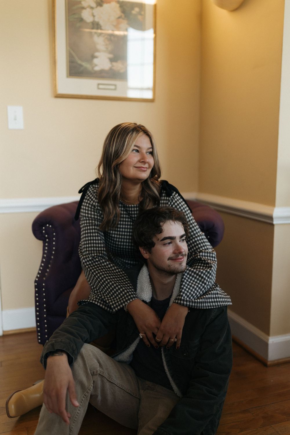 A couple shares intimate moments while sitting on a chair in a room with beige walls and hardwood floors.