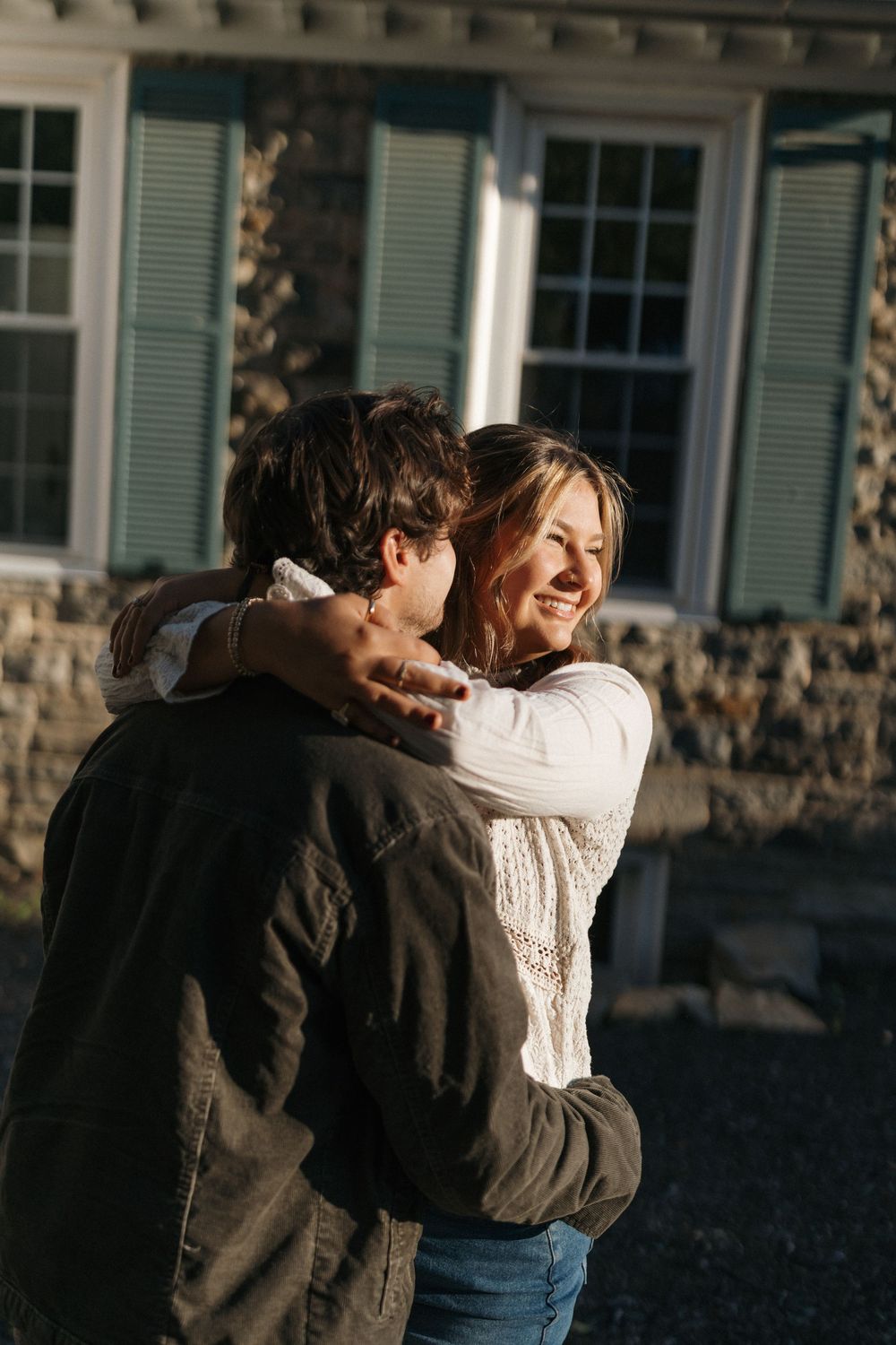 A couple embraces and laughs together in front of a house with green shutters during a sunny day.