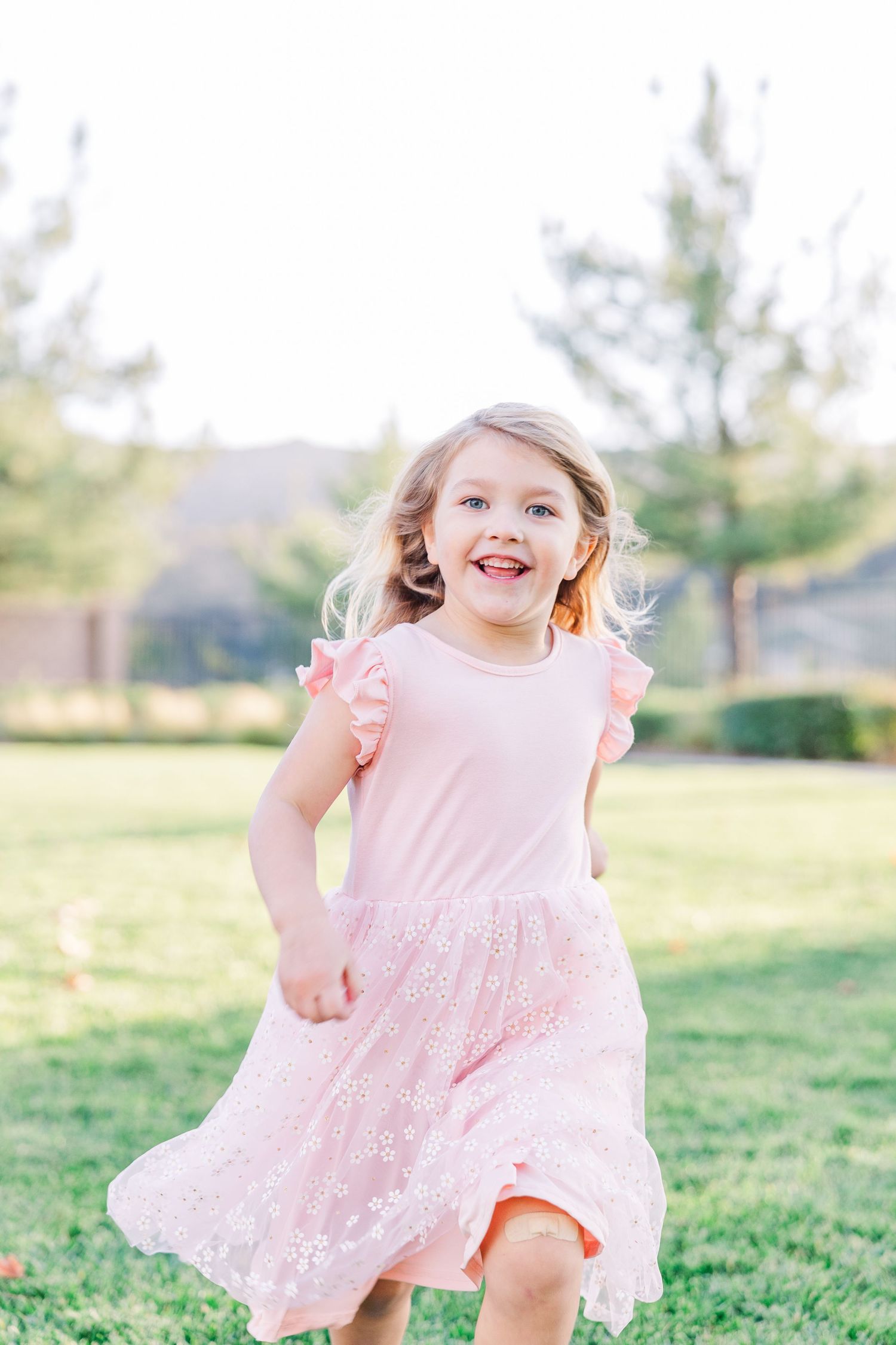 A young child wearing a pink lace dress twirls and plays in a grassy field on a sunny day.