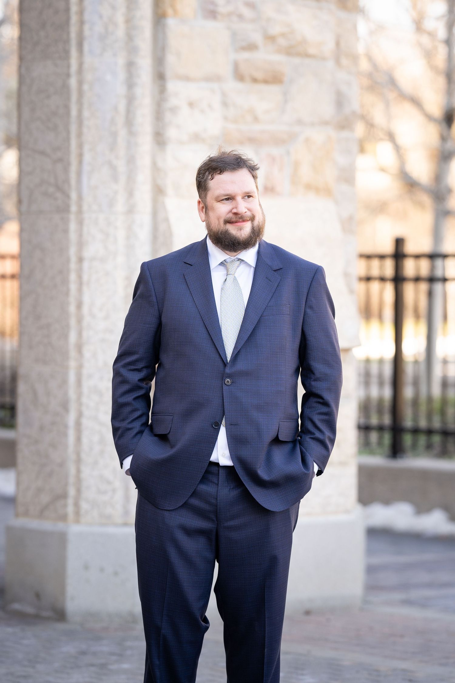 A groom in a navy blue suit and light tie stands with hands in pockets against a stone building exterior.