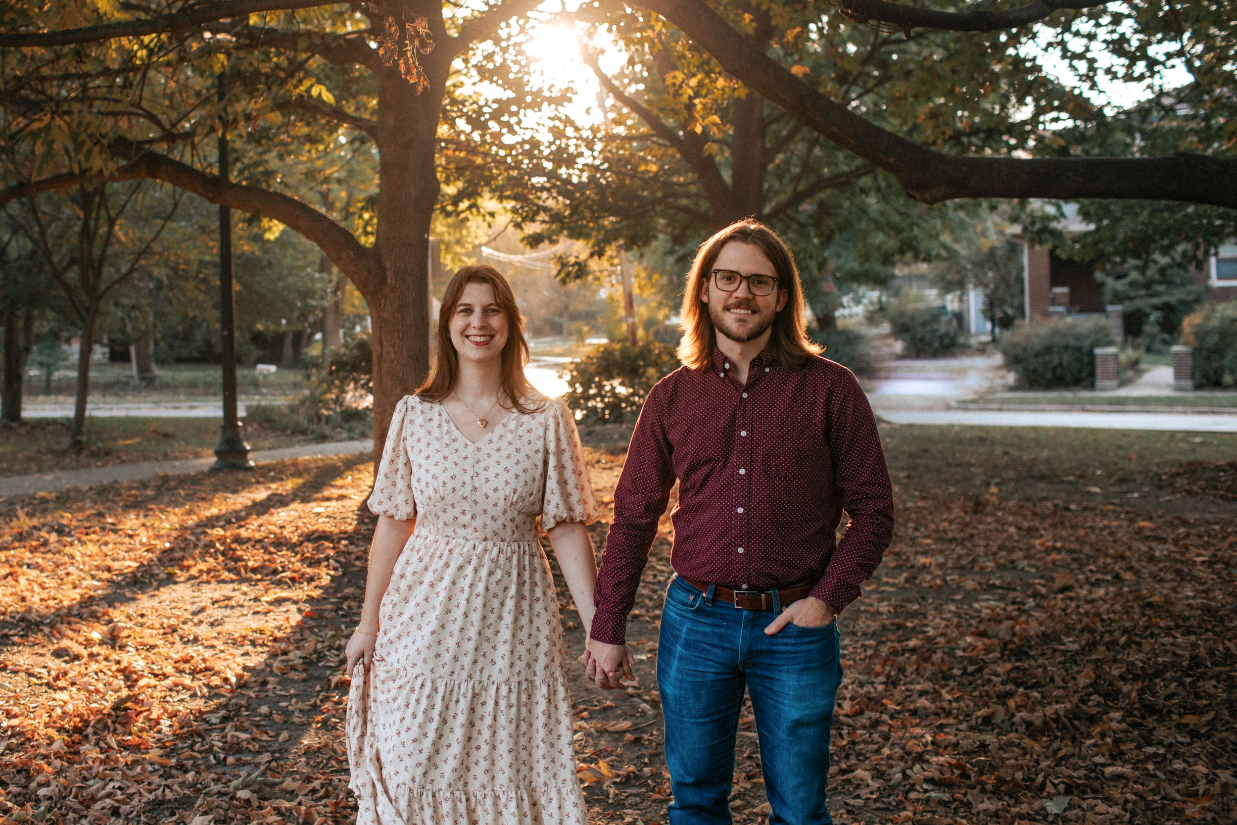 A couple walks together through a park during golden hour as autumn leaves cover the ground and sunlight filters through trees.