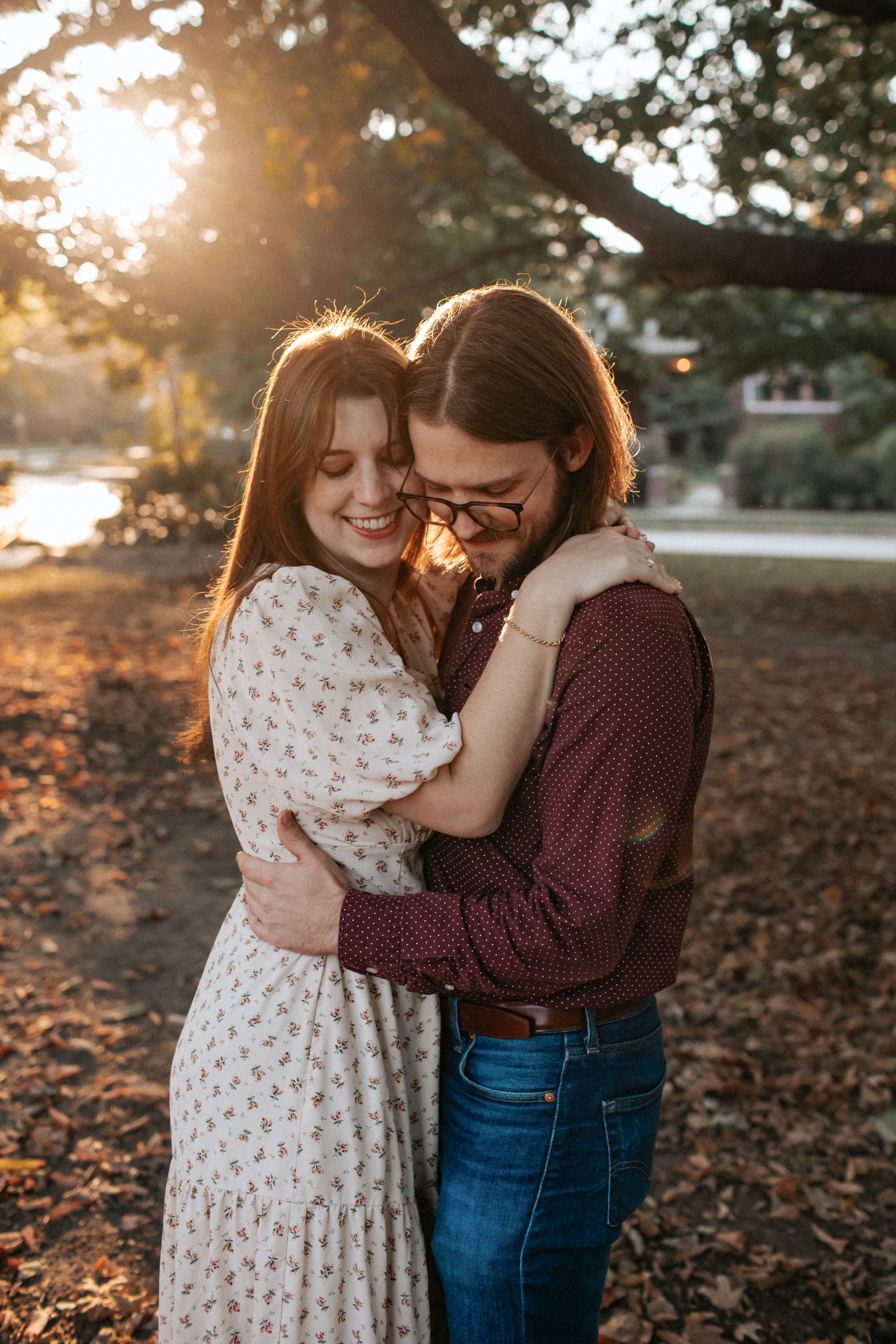 A couple embraces and laughs together during a romantic sunset photoshoot in a park with fallen autumn leaves.