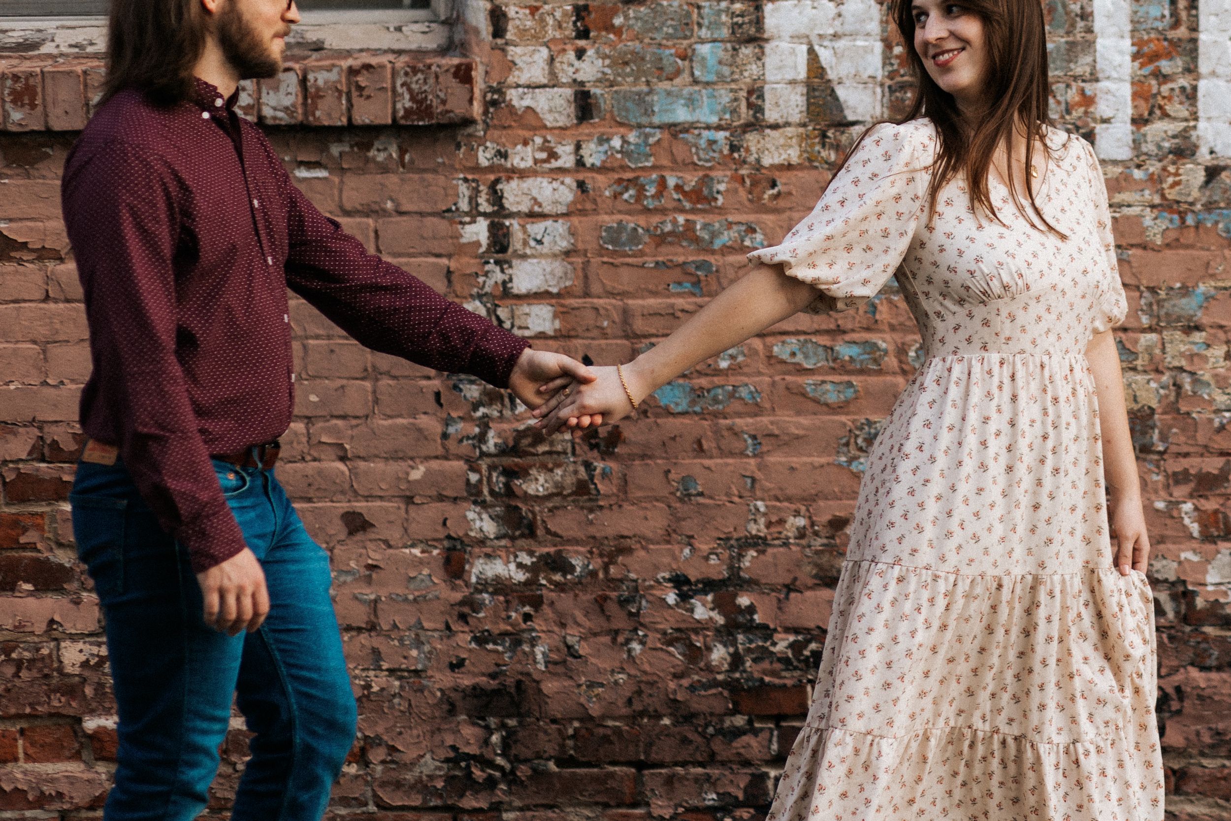 A couple in casual attire poses against a rustic brick wall background in a romantic engagement photo session.