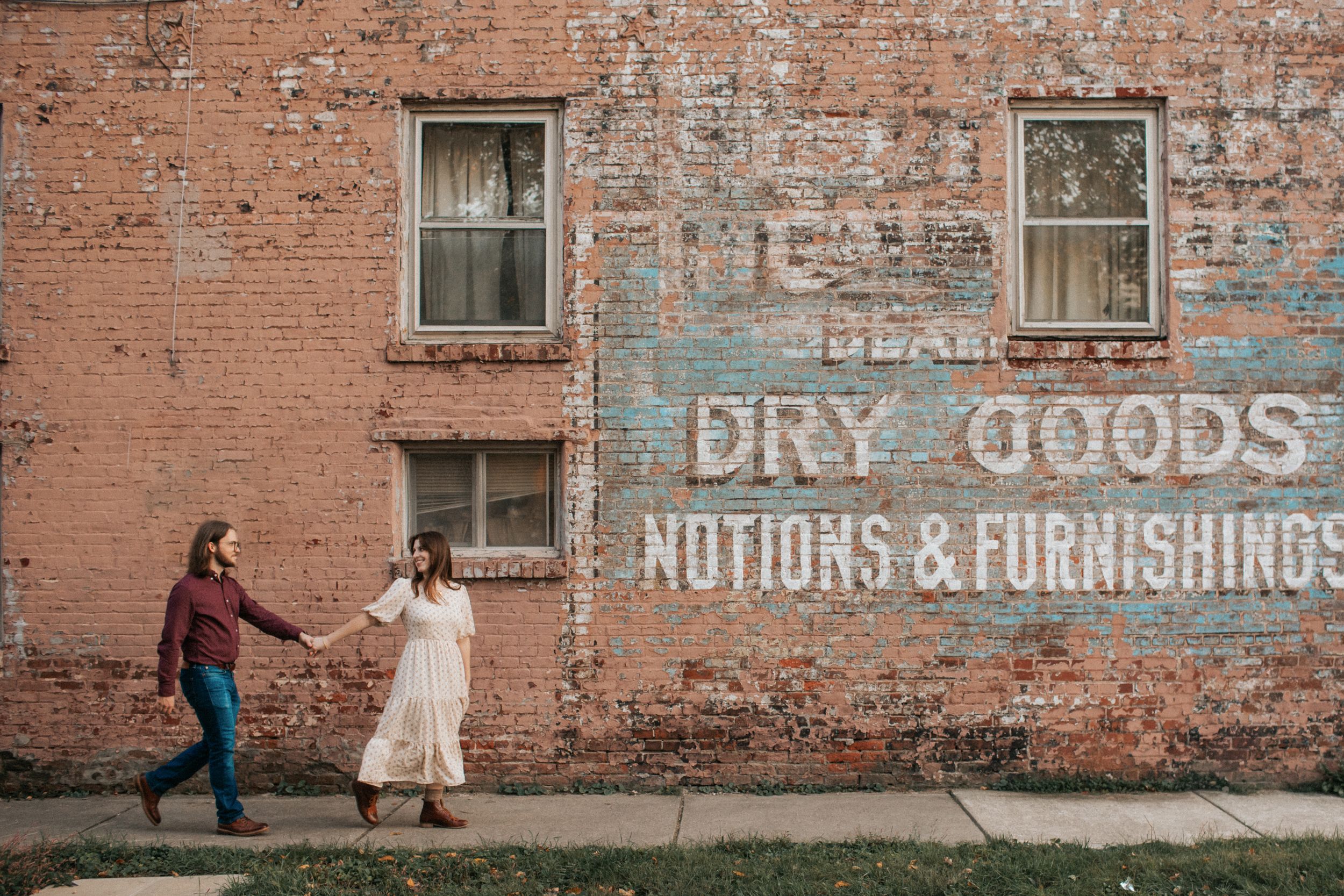People walk past faded vintage signs for dry goods and furnishings painted on weathered brick walls of an old building.