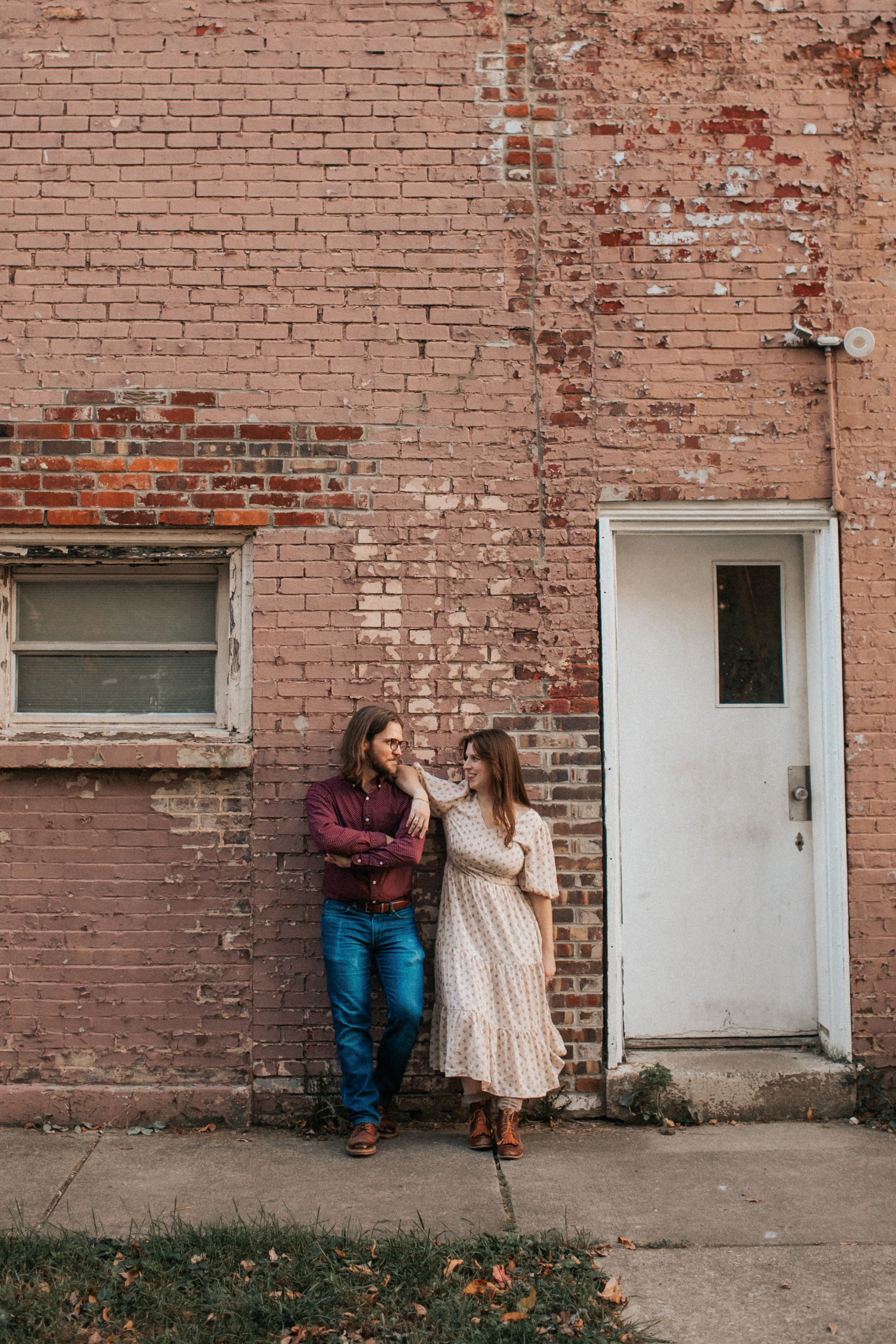 A couple shares a tender moment against a weathered brick wall with a white door and window.