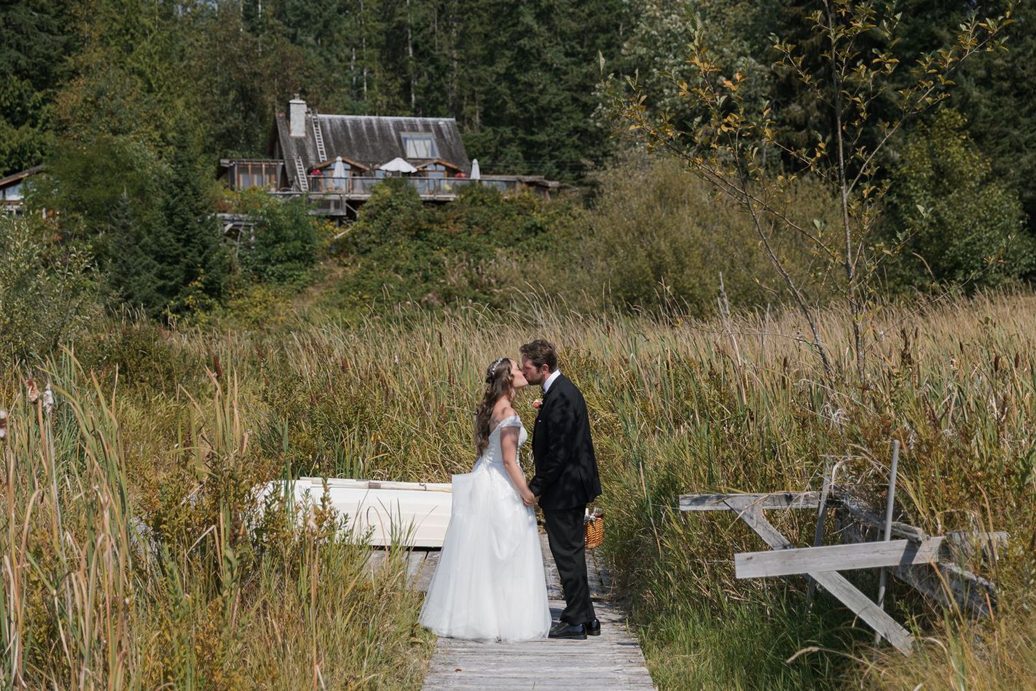 Bride and groom share a kiss on a wooden dock surrounded by tall golden grass and trees.