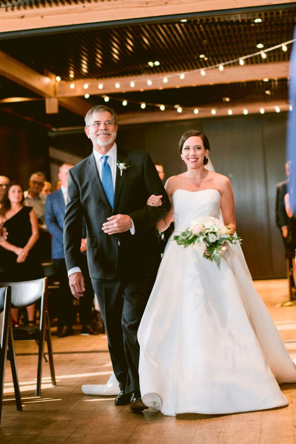 A couple walks down the aisle during a wedding ceremony in an indoor venue with string lights overhead.