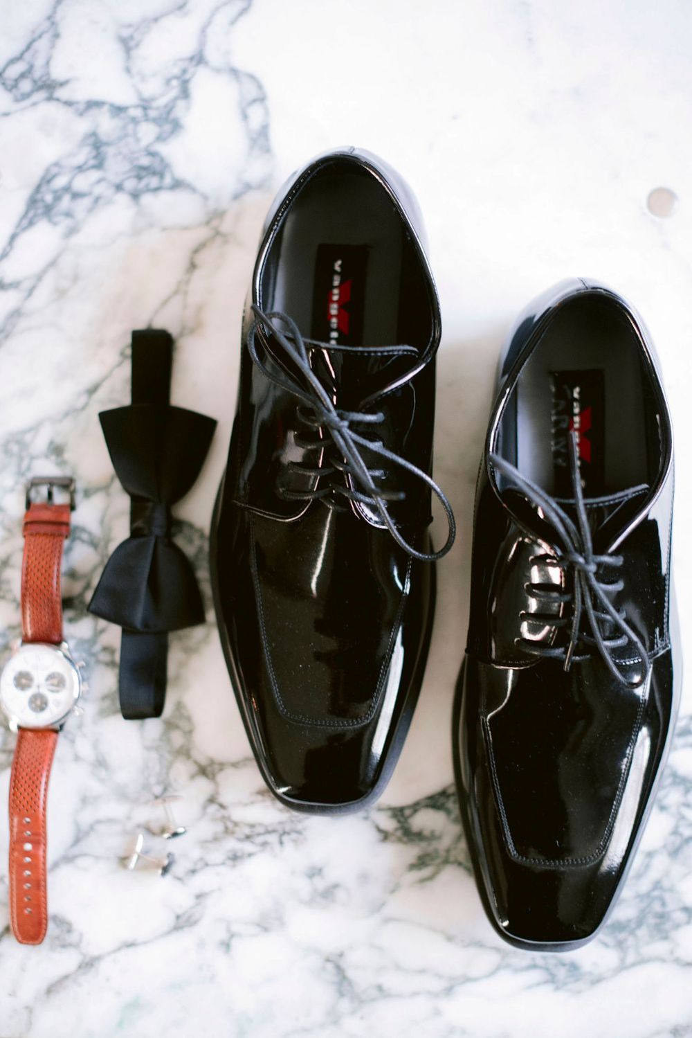 A row of shiny black dress shoes and watch straps arranged on a white marble surface.
