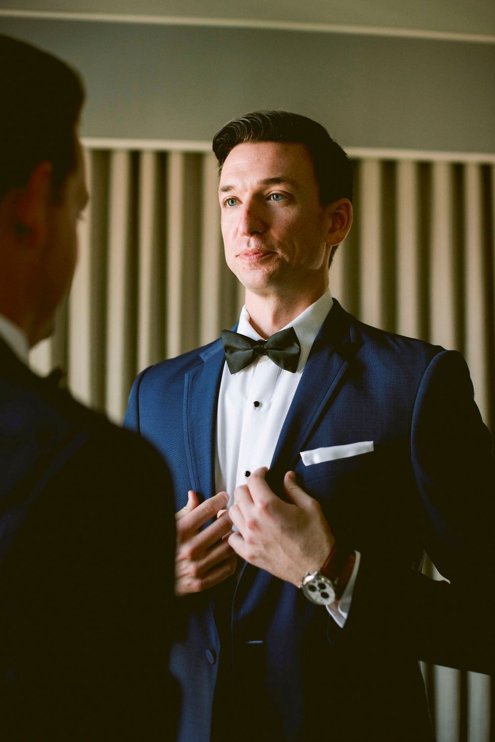 A groom in a navy blue tuxedo adjusts his bowtie while getting ready for a formal event.