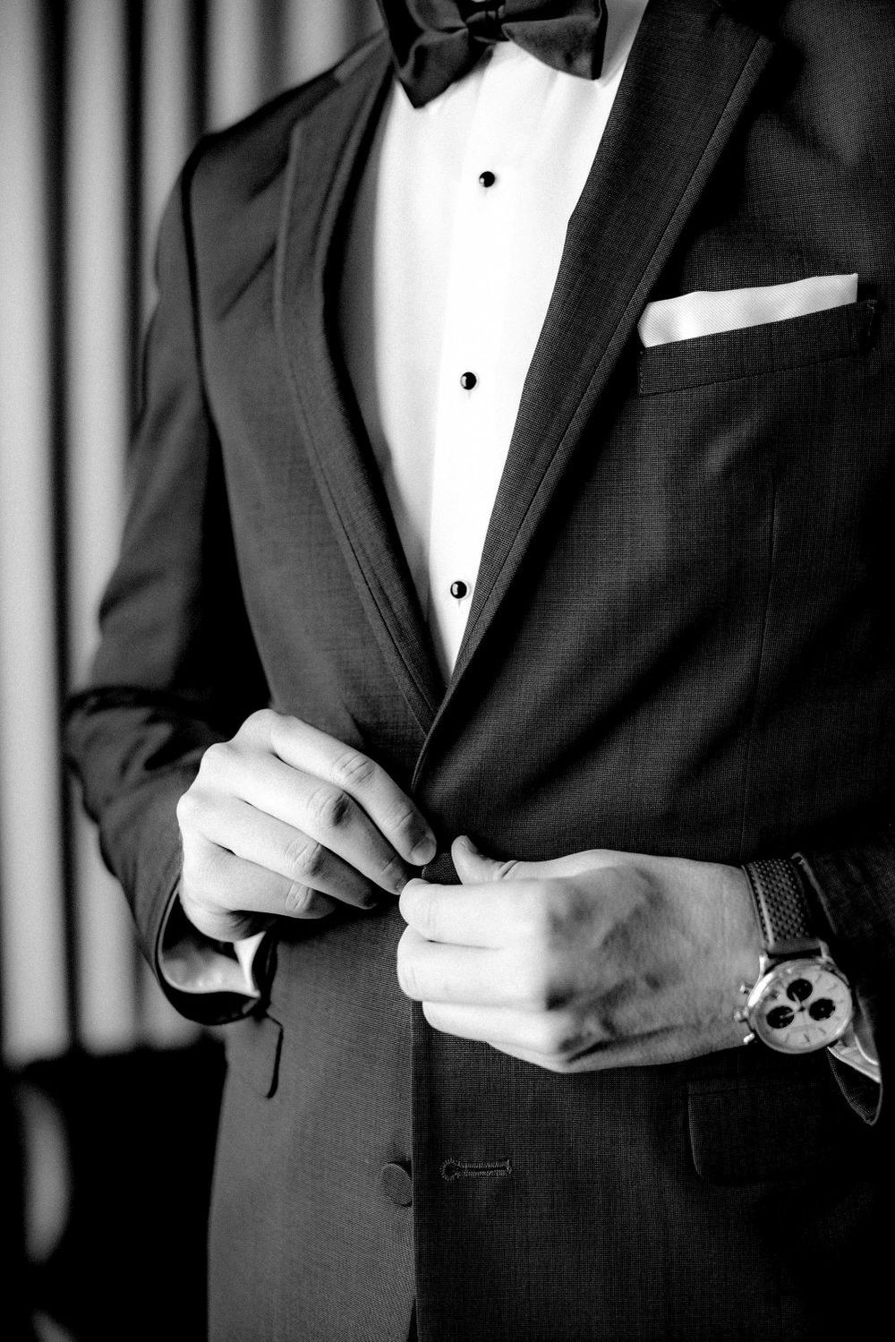 A man in a black tuxedo and bow tie adjusts his cuffs in a formal black and white photograph.