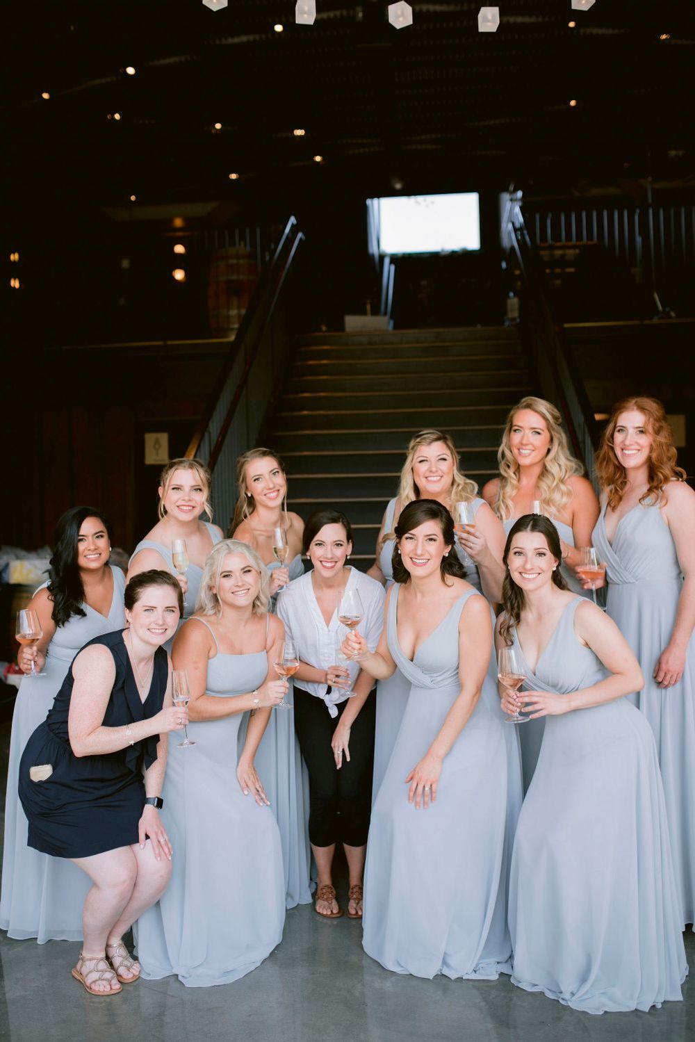 A group wearing light blue bridesmaid dresses gather together on dark stairs for a wedding photo.
