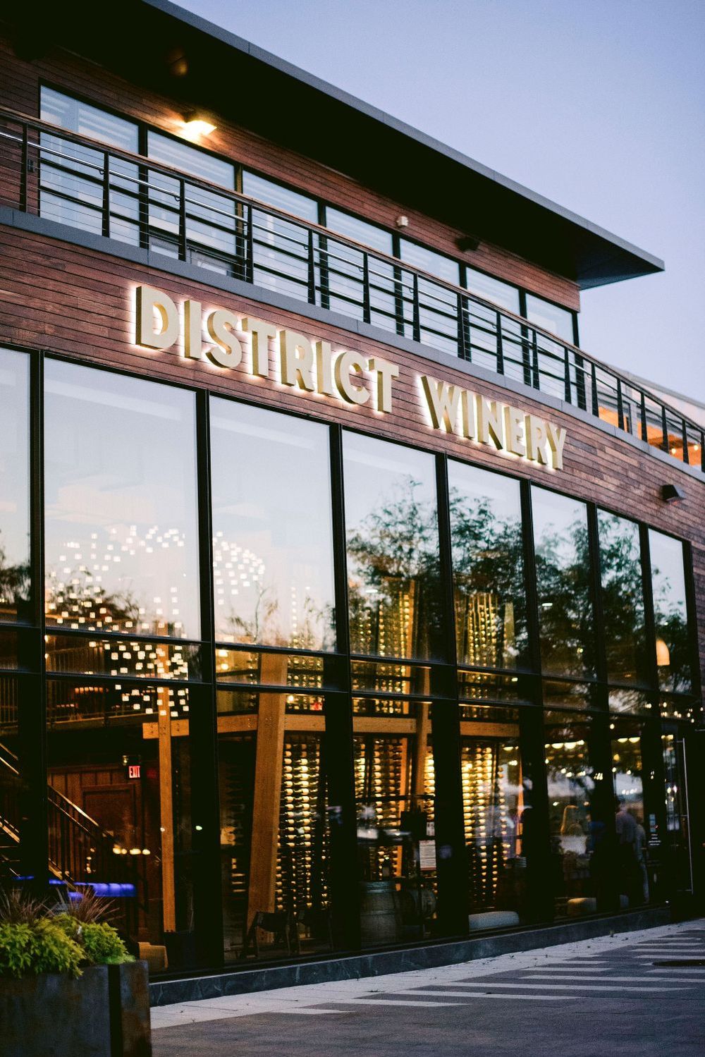 Modern storefront exterior with large glass windows and illuminated 'District Winery' signage at dusk.