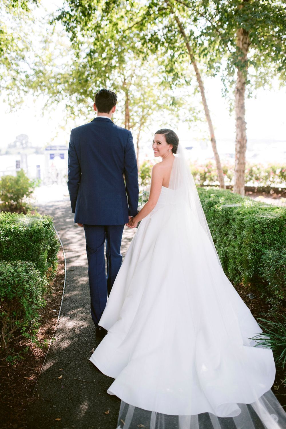 A bride and groom walk along a tree-lined path, with her flowing white wedding dress trailing behind them.