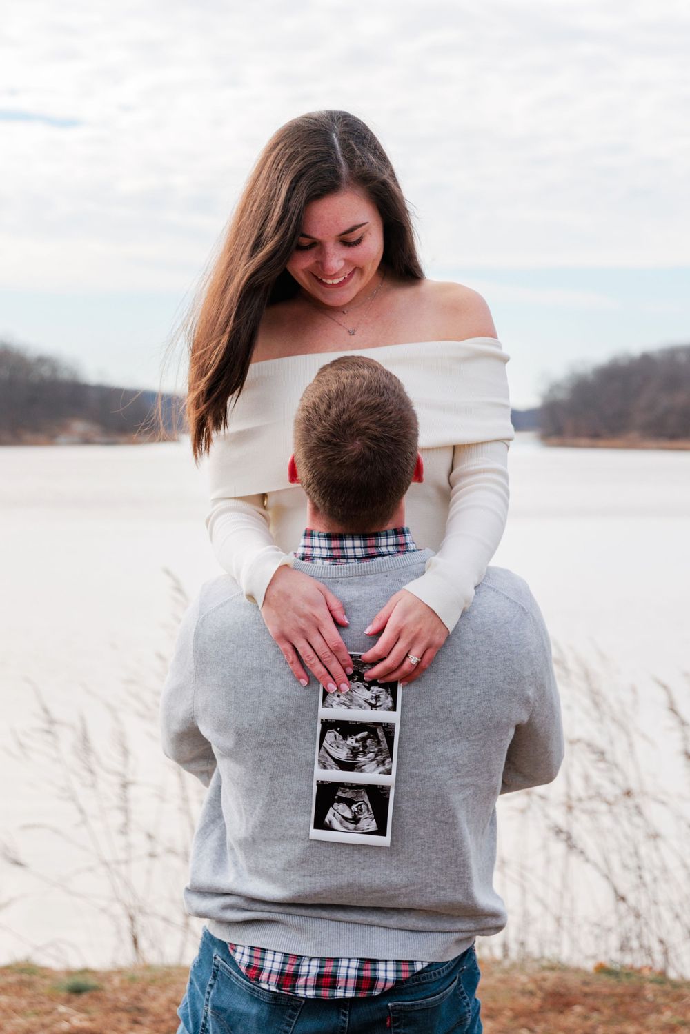 Couple embraces while holding ultrasound photo in outdoor winter setting with tall grass in background.