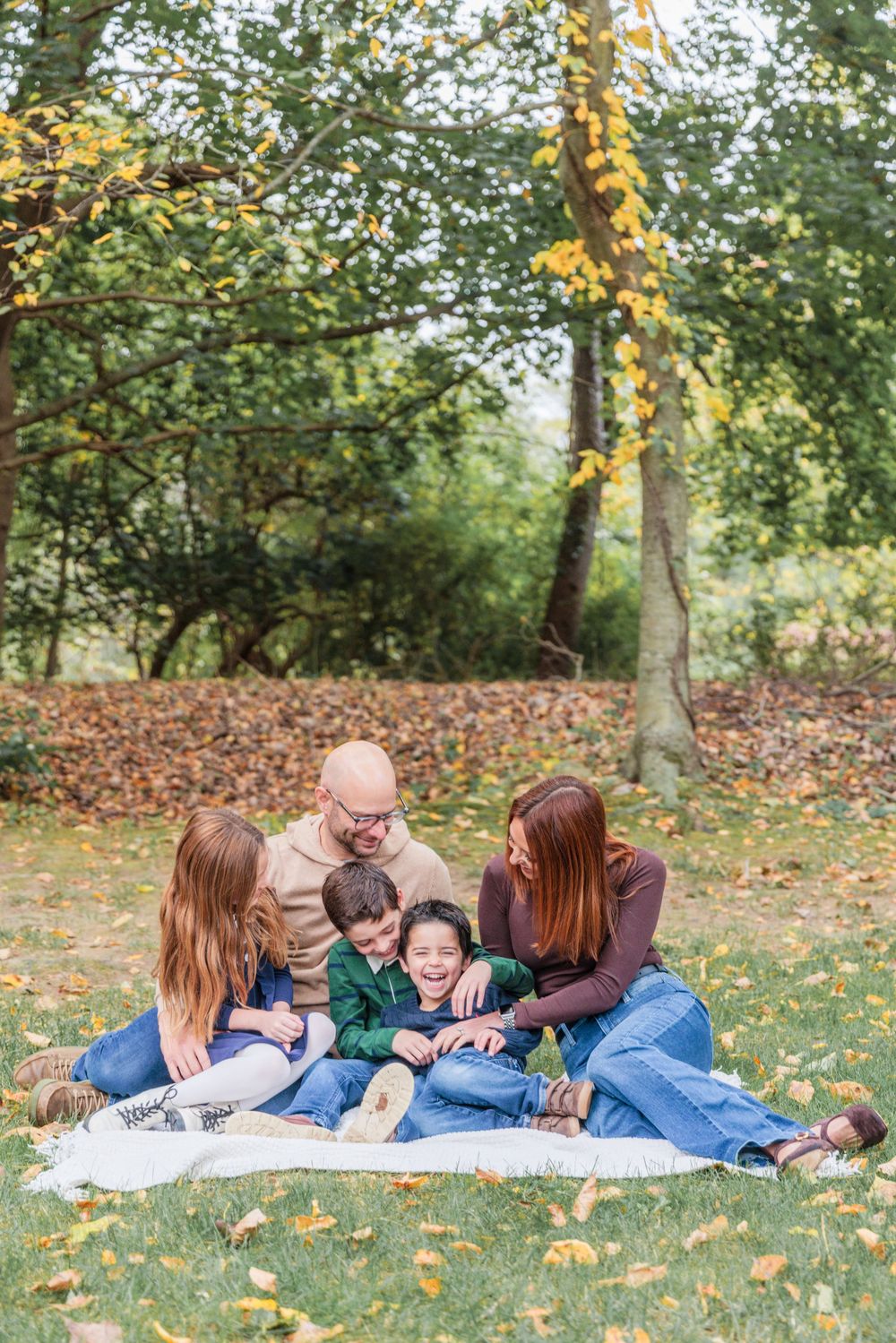 A family lies together on a blanket surrounded by fallen autumn leaves in a park setting.