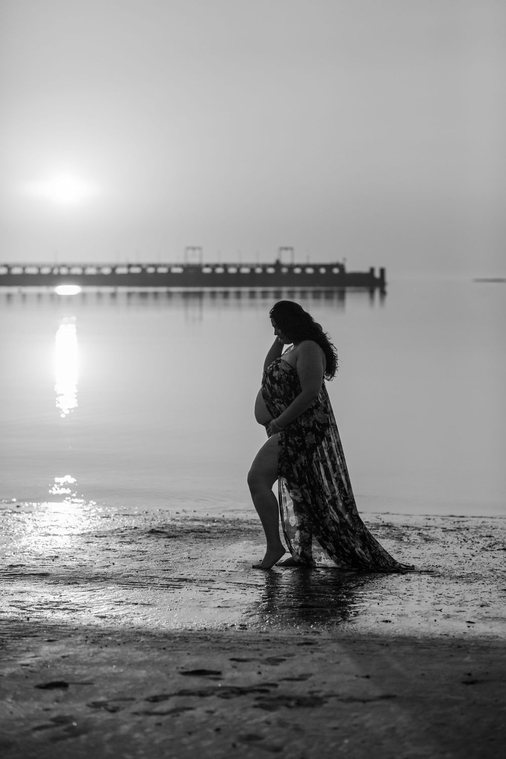 Silhouette of a pregnant woman in flowing dress stands on misty beach at sunset with pier in background.