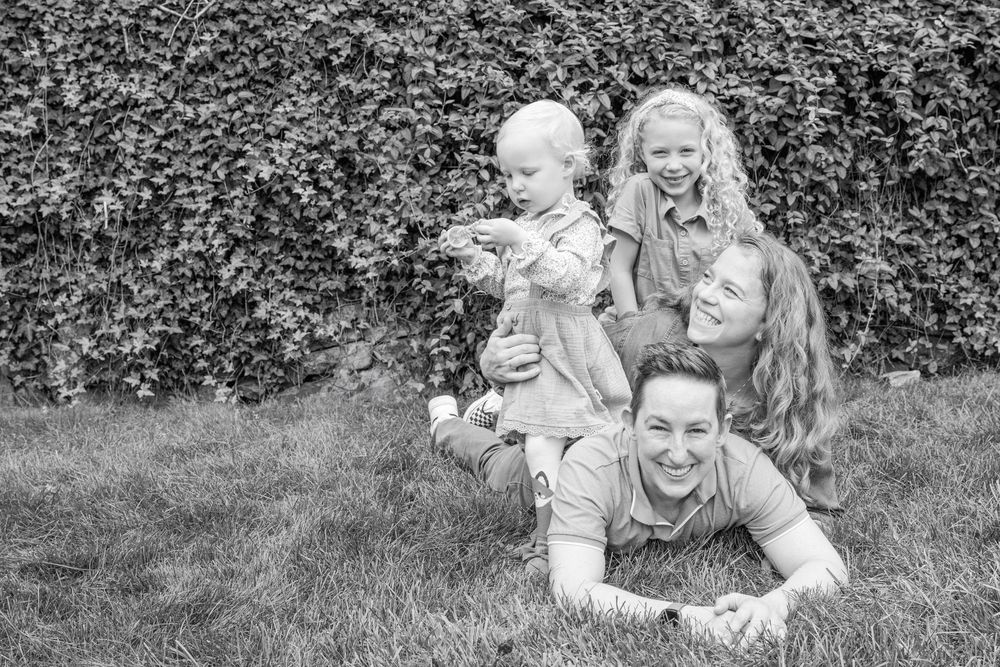 Black and white photo of a Family of 4 laying on the grass. Dad is on the bottom, with mom stacked on top, and their oldest sitting on mom.