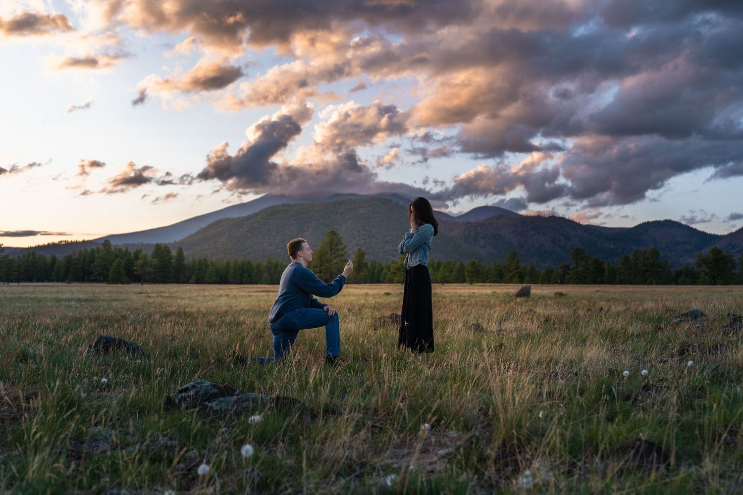 A couple shares a romantic moment in a mountain meadow at sunset with dramatic clouds overhead.
