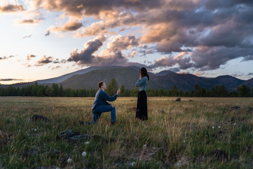 A couple shares a romantic moment in a mountain meadow at sunset with dramatic clouds overhead.