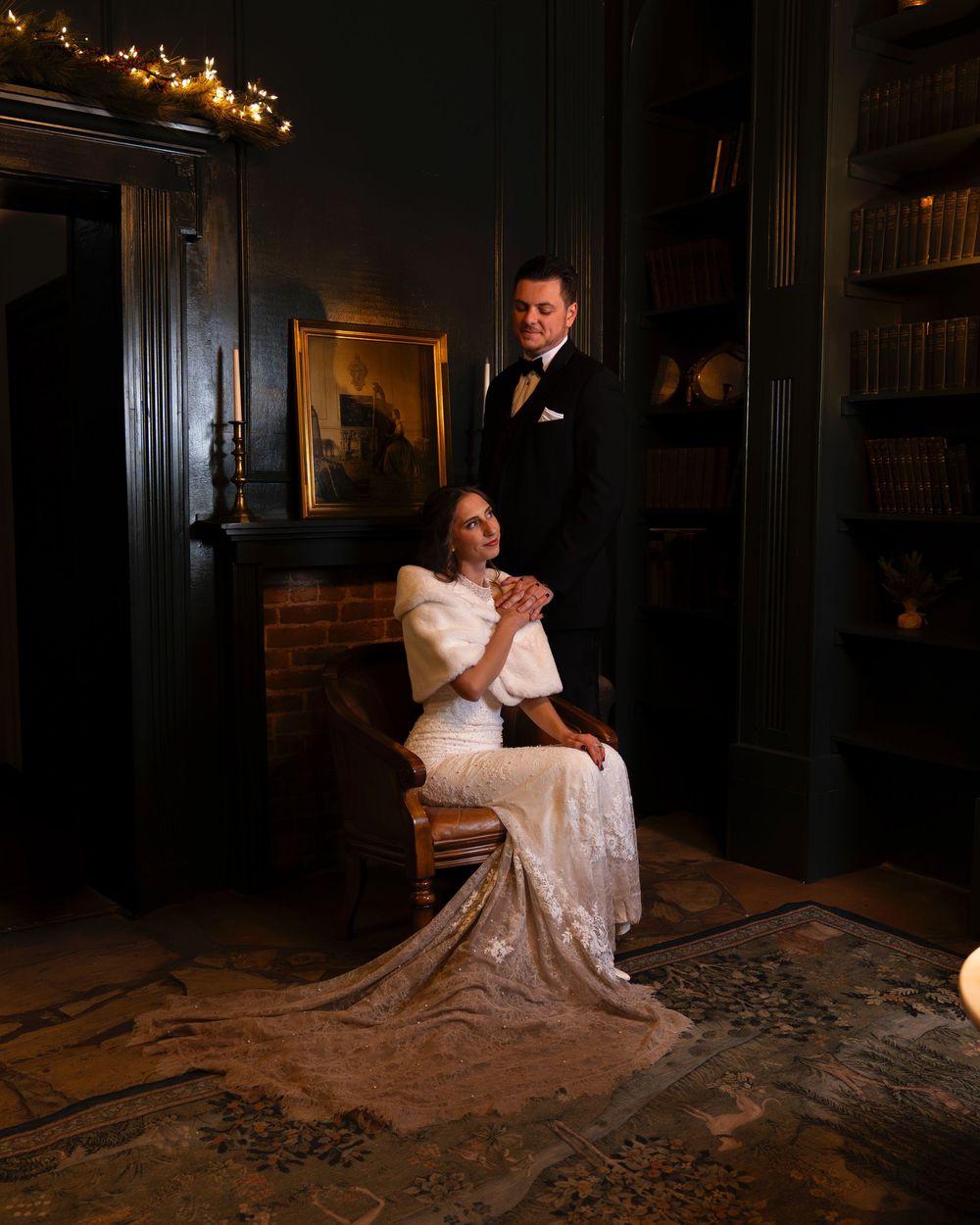 Elegant couple in formal attire posing in dimly lit vintage room with string lights.