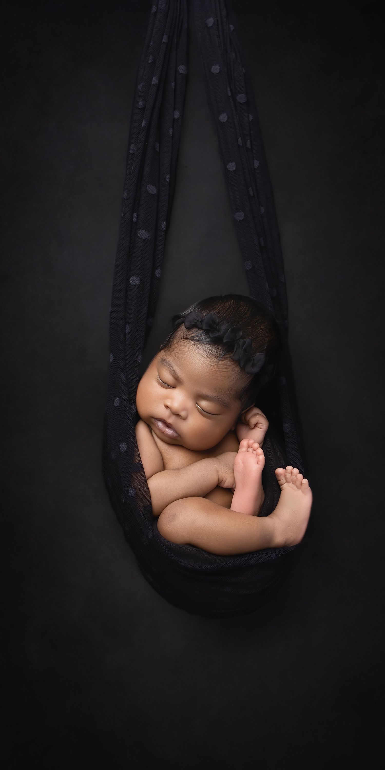 Newborn portrait featuring sleeping baby in a suspended hammock pose posed on a black background