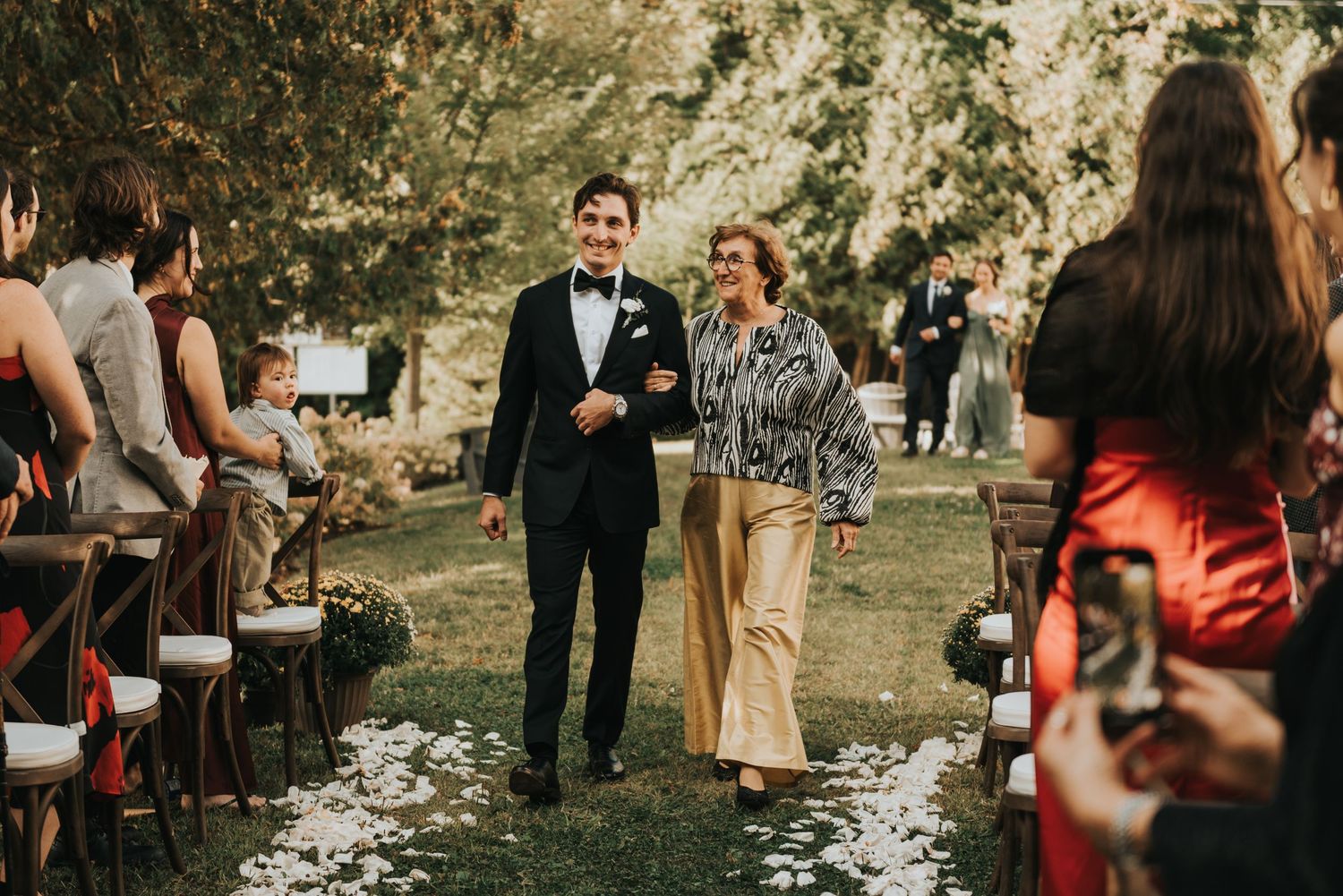 Couple walks down outdoor wedding aisle with scattered white petals while guests watch from wooden chairs.
