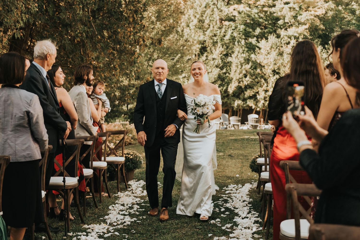 Guests seated at an outdoor wedding ceremony in a lush garden setting with natural decor and white chairs.
