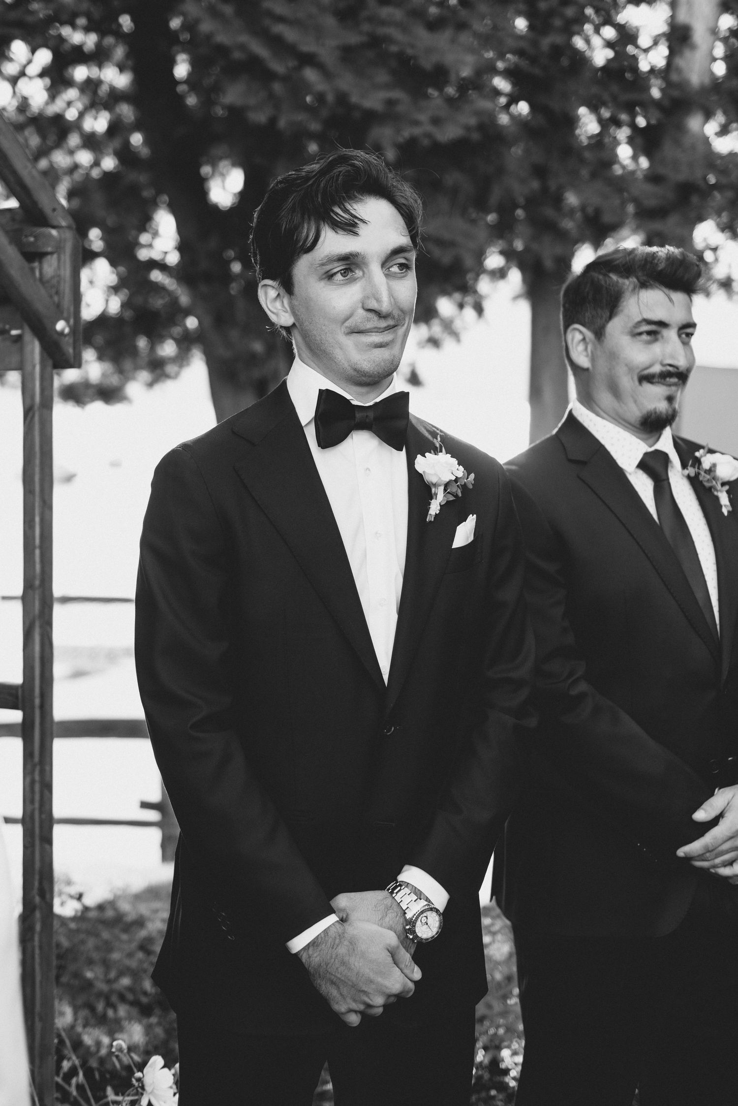 A group of groomsmen in black tuxedos share a candid moment during a wedding celebration outdoors.
