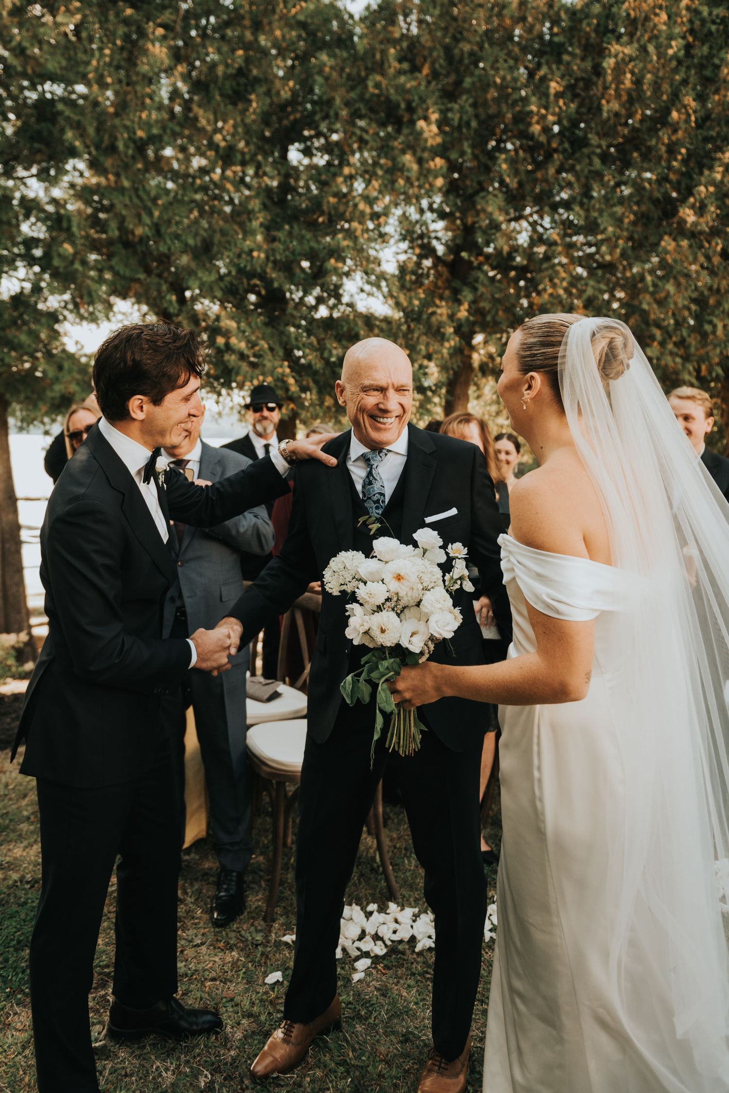 A wedding party gathers outdoors under autumn trees during an emotional ceremony moment.