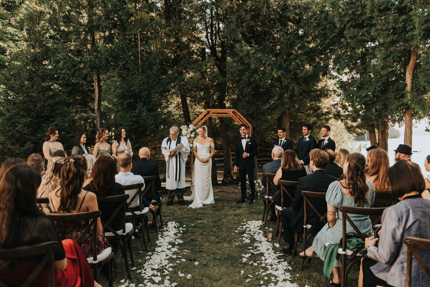 Wedding guests gather outdoors for a ceremony in a wooded setting with natural lighting and rustic decor.