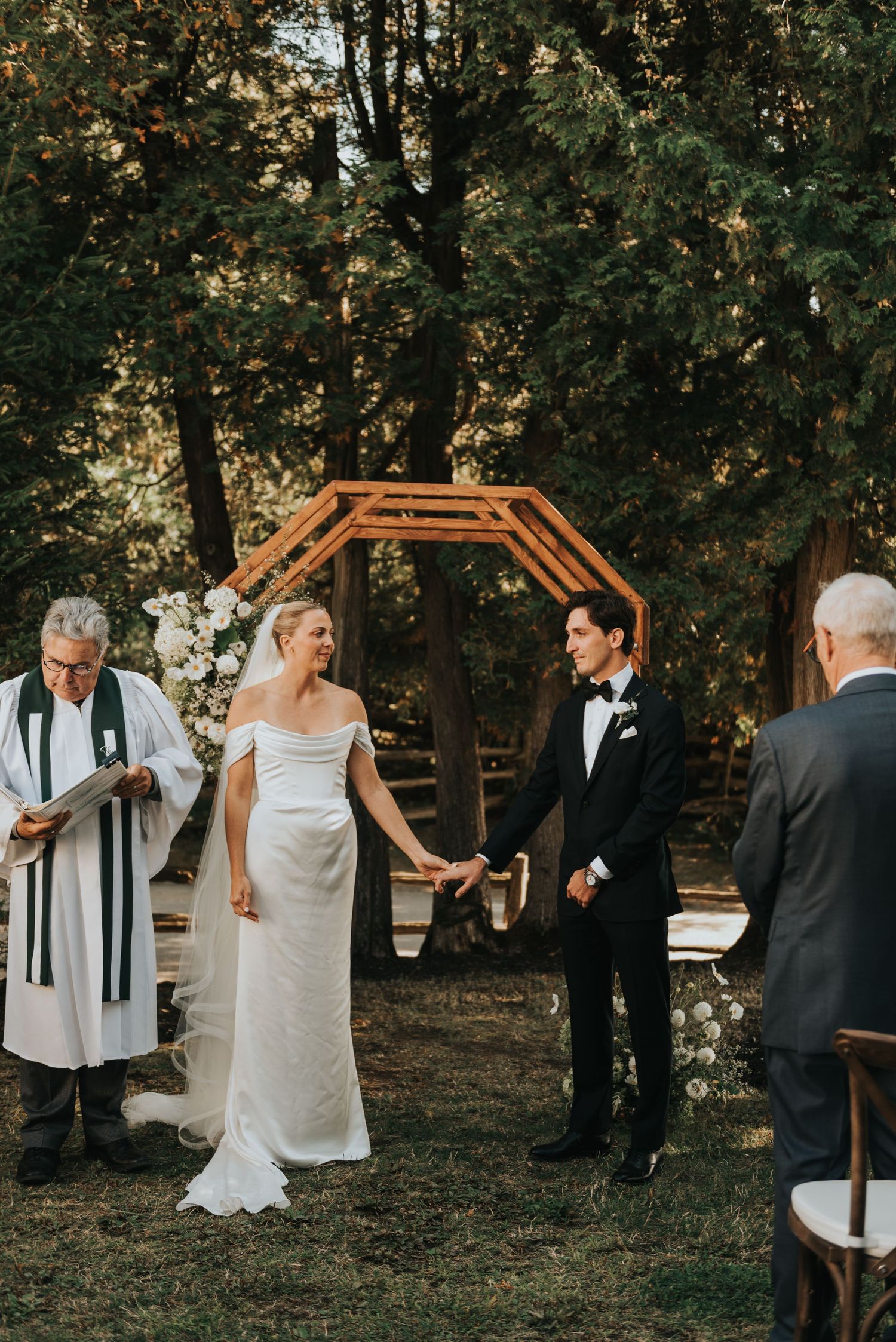 A wedding ceremony takes place under a wooden hexagonal arch in a wooded outdoor setting.