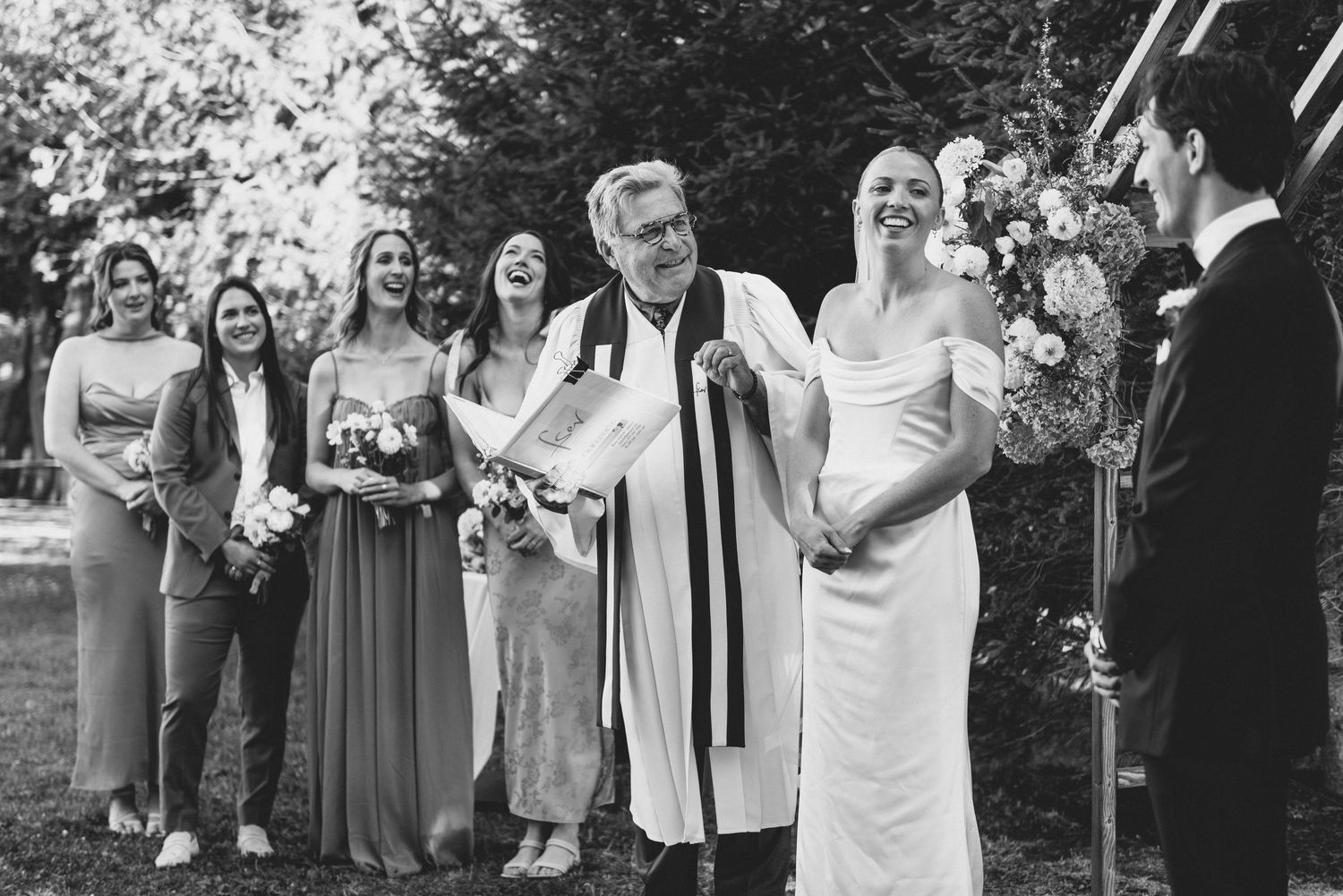 A joyful outdoor wedding ceremony with bridesmaids in flowing dresses and an officiant in white robes.