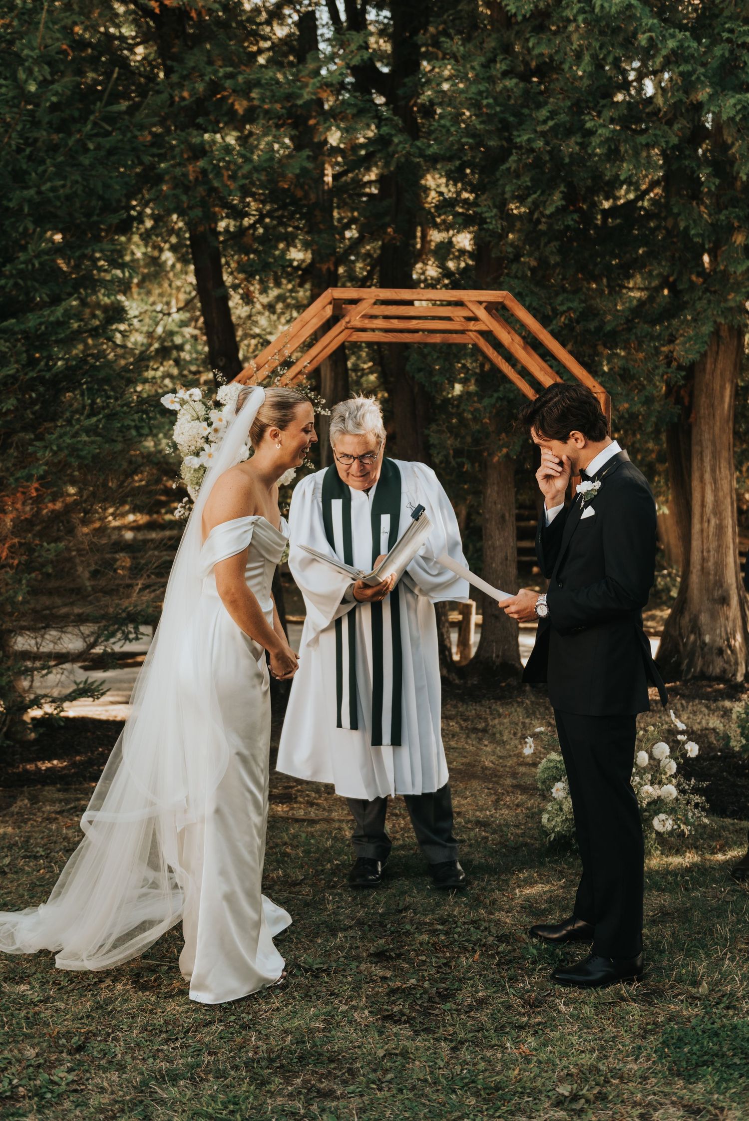 A wedding couple shares joyful moments with clergy in a wooded outdoor setting during their ceremony.
