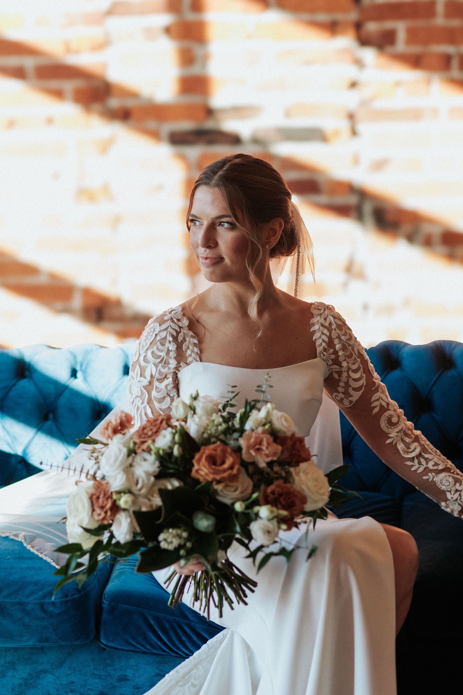 White wedding gown with lace sleeves displayed on blue velvet couch with peach floral bouquet.