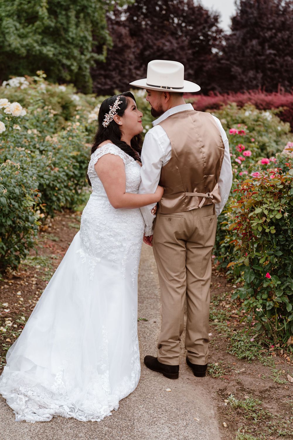 Bride in white lace gown and groom in tan vest and cowboy hat share a romantic moment in a rose garden.