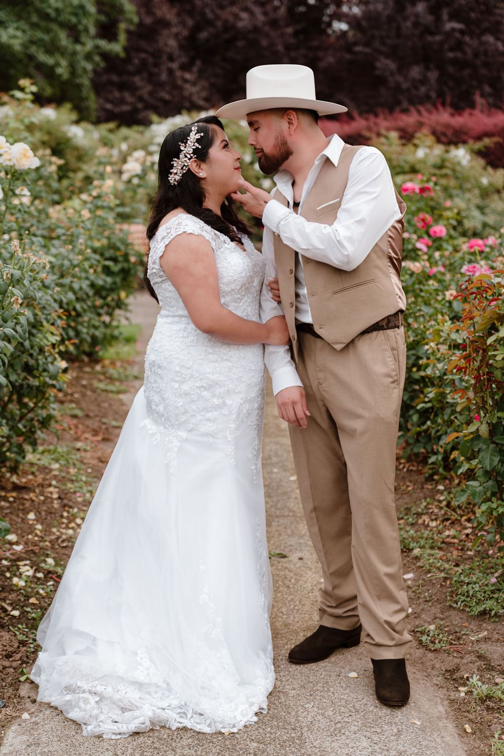 Bride in white lace gown and groom in tan suit with cowboy hat share a romantic moment in a blooming rose garden.