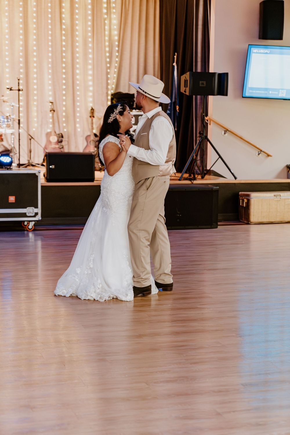 Bride and groom share their first dance on a wooden floor, with a lit stage and guitars in the background.