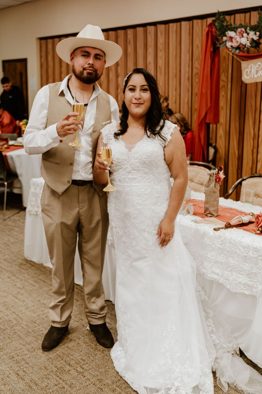 A bride in white lace gown and groom in tan suit with white hat toast with champagne at their wedding reception.