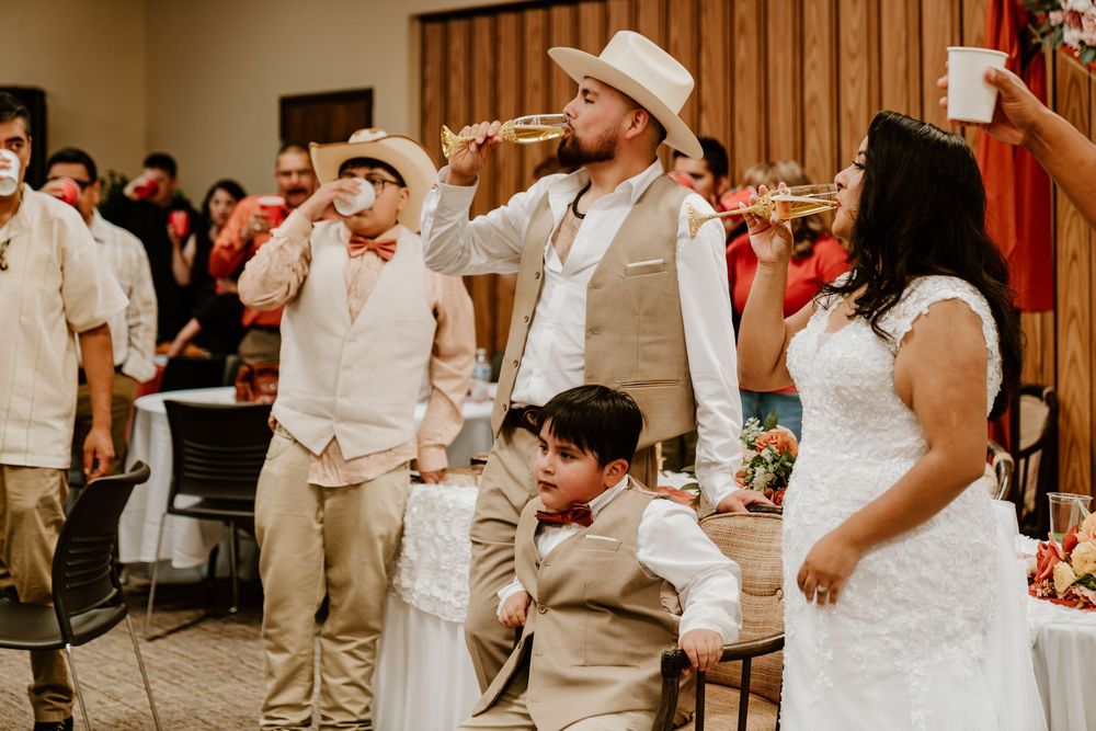 Wedding party in tan suits and white hats celebrate indoors, with a trumpet player and bride in white dress.