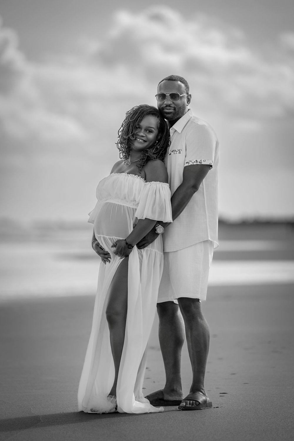 Black and white beach maternity photos showing intimate couple poses against cloudy ocean backdrop.